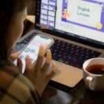 Adult woman multitasking, using laptop and smartphone while studying English with tea on a table.