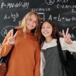 Two smiling students with backpacks in front of a math chalkboard in a classroom.
