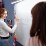 Smiling teacher writing on a whiteboard in a lively classroom setting with students.